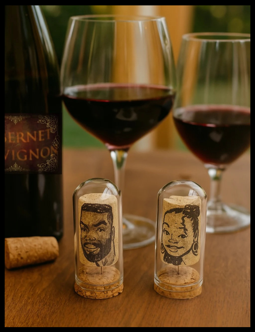 Wine bottle, glasses, and cork art with faces on a wooden surface.